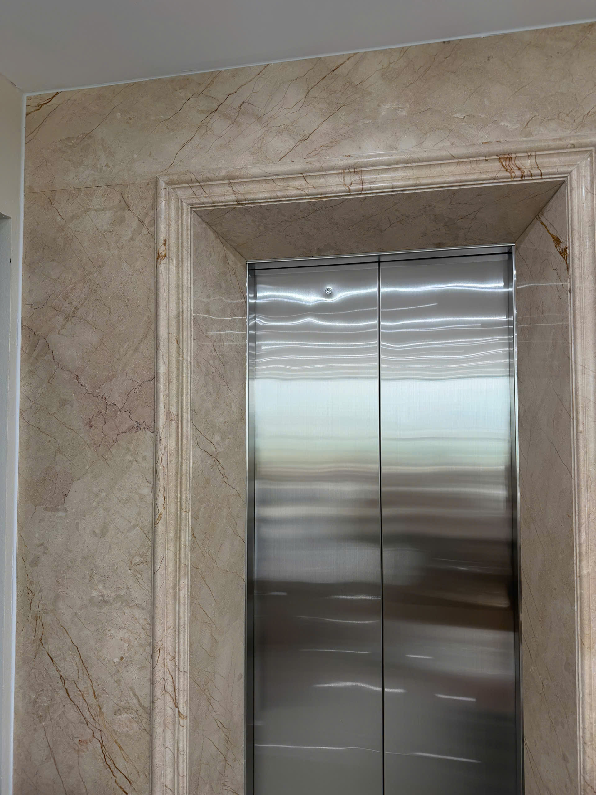 Cửa thang máy inox đóng kín, khung ốp đá hoa cương màu be trong sảnh tòa nhà